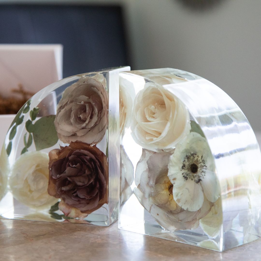 A pair of arch-shaped clear resin bookends with preserved florals including roses and anemones in soft cream, blush, and mocha tones, displayed on a light surface.