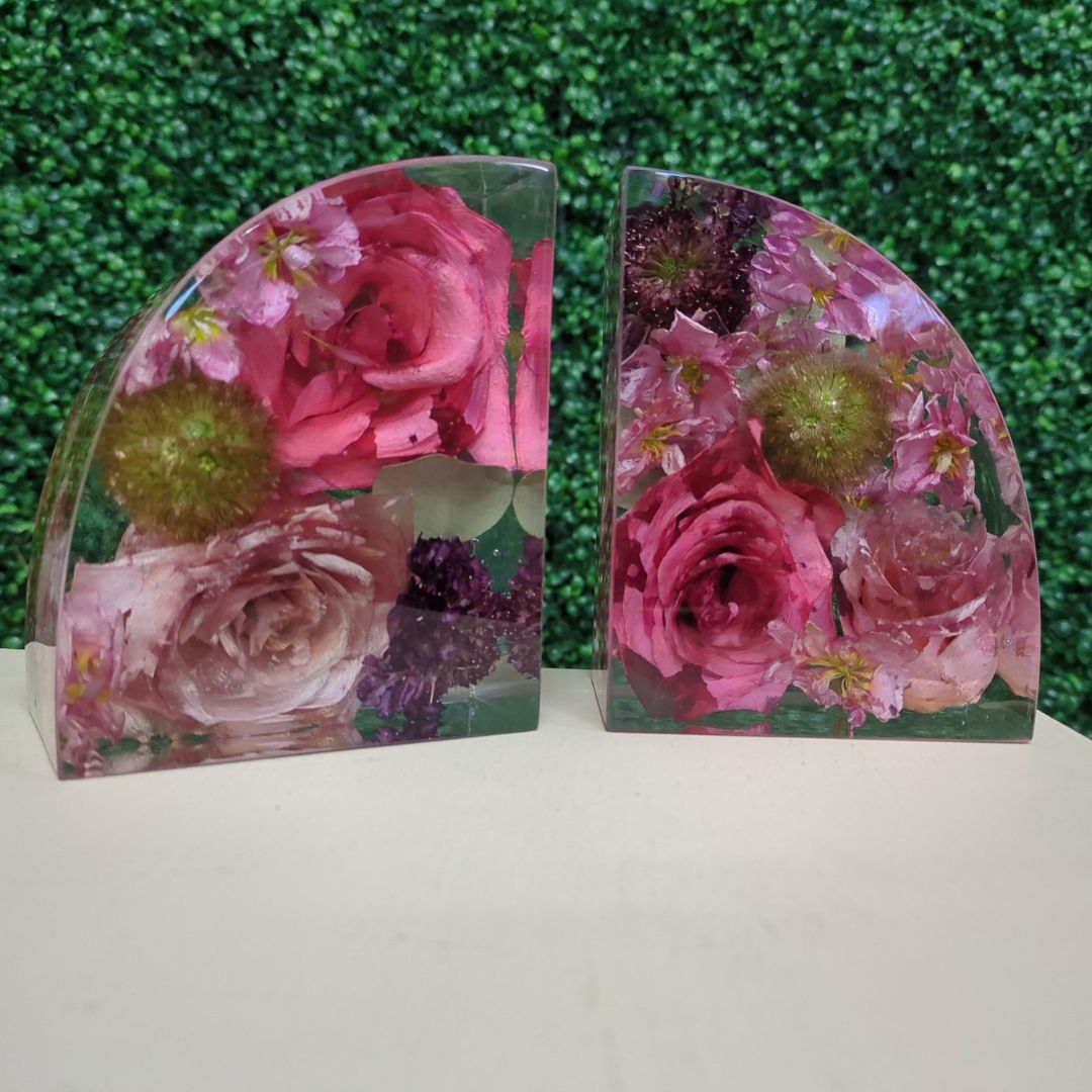 A pair of arch-shaped clear resin bookends filled with preserved florals including vibrant pink roses, green button mums, and mixed blooms, displayed on a white surface with a green hedge wall in the background.