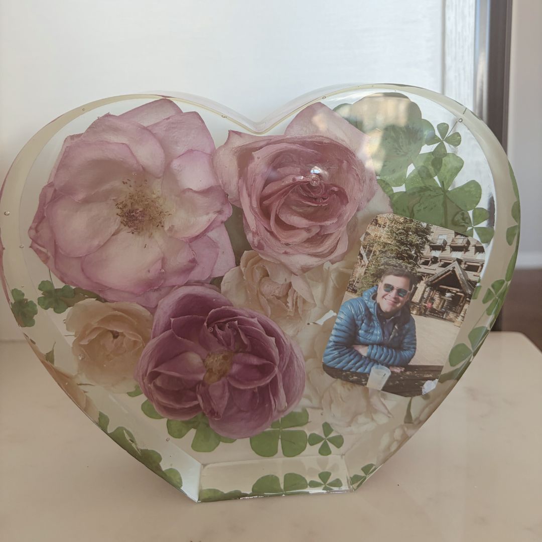 A heart-shaped clear resin keepsake filled with preserved florals including pink and cream roses, small green leaves, and an embedded personal photo, displayed on a light surface.