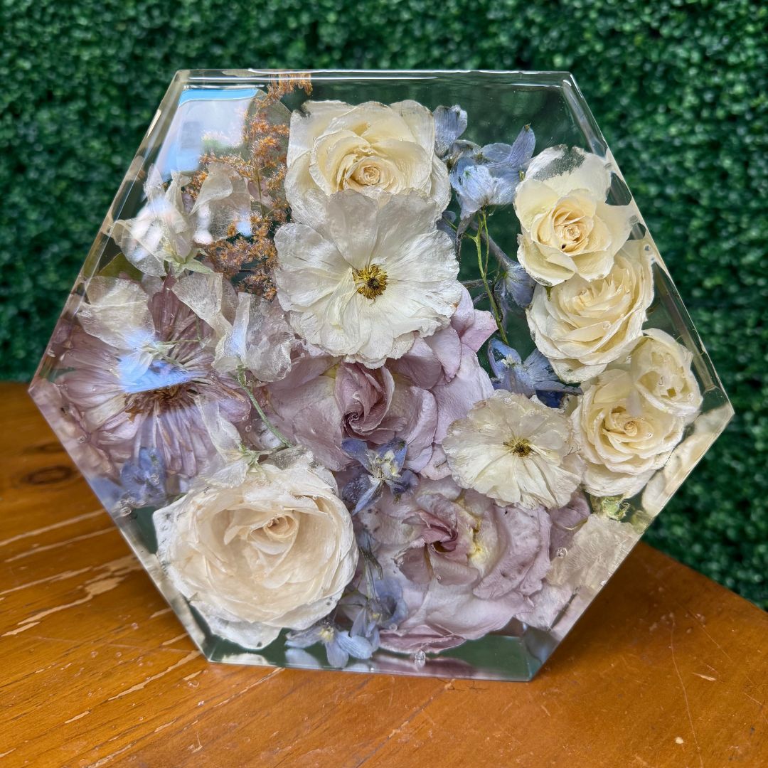 A hexagon-shaped clear resin keepsake filled with preserved wedding florals including roses, hydrangeas, and delicate blooms in soft cream, blush, and lavender tones, displayed on a wooden surface with a green hedge wall in the background